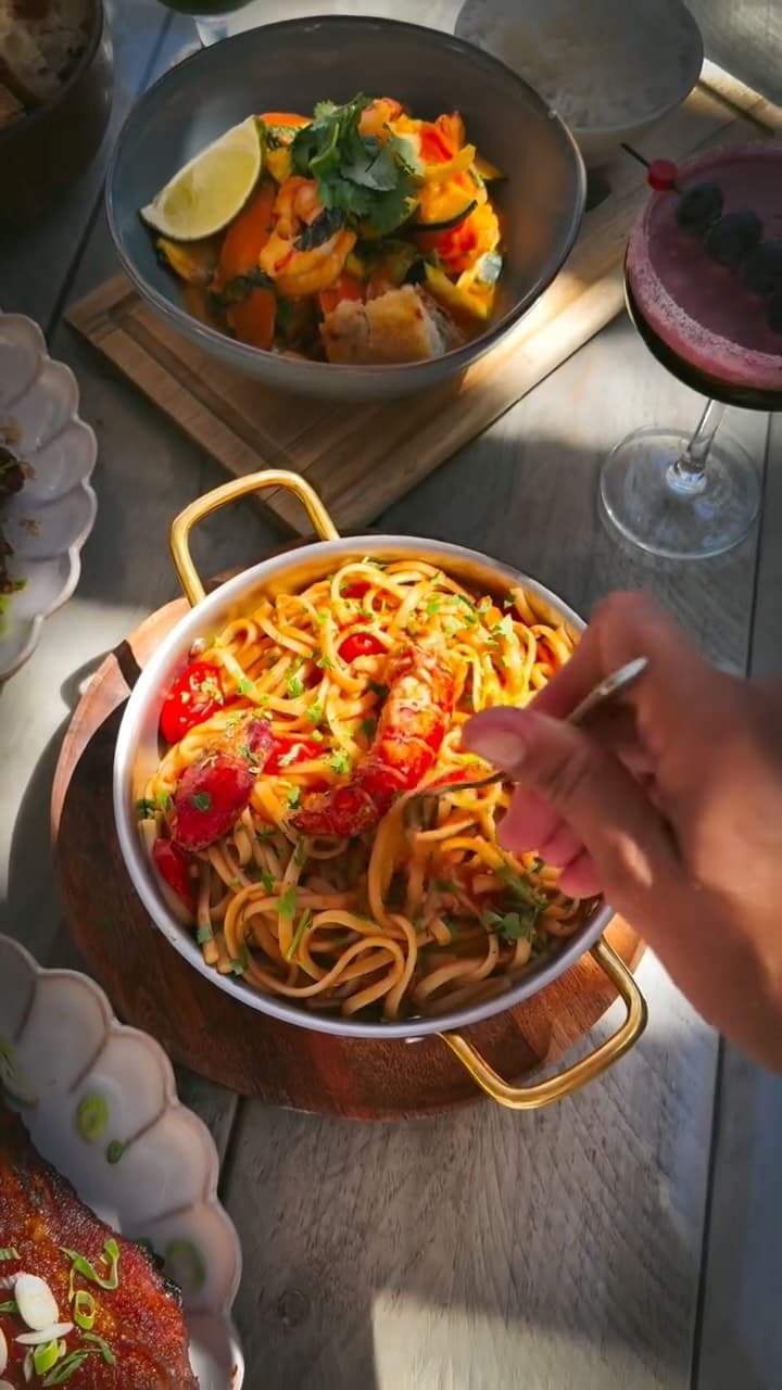 A hand uses a fork to eat lobster pasta served in a metal pan with gold handles, accompanied by a bowl of shrimp and vegetable stir-fry with lime and cilantro, a glass of purple cocktail garnished with blueberries, and a side of rice on a wooden table.